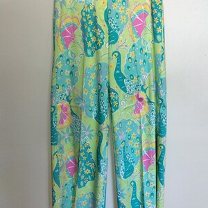 May include: Wide-leg trousers featuring a vibrant, multi-coloured peacock and floral pattern. The design includes shades of lime green, teal, yellow, and pink, with stylised peacock figures and small yellow flowers printed across the entire fabric.