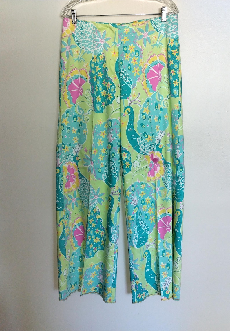 May include: Wide-leg trousers featuring a vibrant, multi-coloured peacock and floral pattern. The design includes shades of lime green, teal, yellow, and pink, with stylised peacock figures and small yellow flowers printed across the entire fabric.