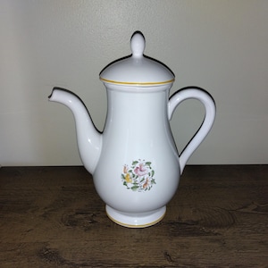 May include: White ceramic coffee pot with a lid and handle. The pot features a floral design and yellow trim. The spout is on the left side. The pot is sitting on a wooden surface.