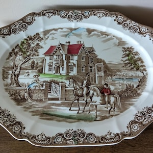 May include: An oval ceramic serving platter featuring a detailed transferware scene of a large house with a red roof, two people on horseback, and a landscape background. The rim is decorated with a brown and white floral and scroll pattern.
