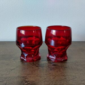 May include: Two ruby red vintage drinking glasses. The glasses have a faceted design on the lower portion and a smooth rim. They are sitting on a dark wooden surface. The glasses are empty.