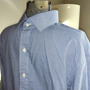 May include: A light blue dress shirt with a small white checkered pattern. The shirt has a classic collar and button-down front. The long sleeves are visible, and the shirt is displayed on a mannequin.