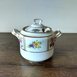May include: White porcelain sugar bowl with a lid and a small spoon. The bowl has a floral design with pink, purple, yellow, and green flowers. The lid and the handle are gold.