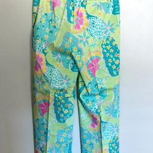 May include: A pair of trousers featuring a vibrant, multi-coloured print on a light green background. The pattern includes stylised peacock motifs in shades of turquoise, bright pink, and yellow, with small floral accents throughout the design.