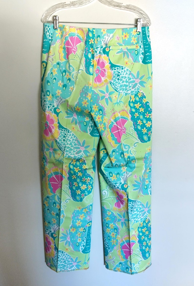 May include: A pair of trousers featuring a vibrant, multi-coloured print on a light green background. The pattern includes stylised peacock motifs in shades of turquoise, bright pink, and yellow, with small floral accents throughout the design.