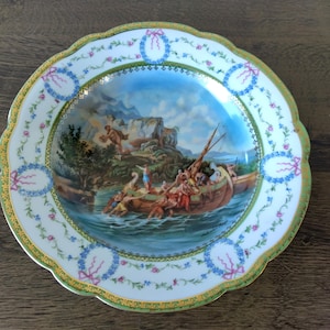 May include: Decorative porcelain plate with a scalloped edge. The plate features a central scene of people in a boat on water, surrounded by a border of floral and ribbon designs. The rim has a gold trim.