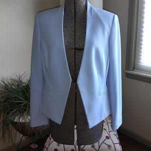 May include: A light blue blazer featuring a collarless design with a deep V-neckline, long sleeves, and two horizontal welt pockets on the front. The jacket is structured with clean lines and a smooth finish, displayed on a dress form.