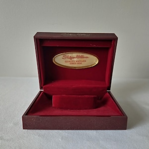 May include: A burgundy watch box with a red velvet interior. The box has a gold-colored oval plaque that reads "Shifrin-Willens QUALITY WATCHES SINCE 1926". The box is open, revealing a watch holder.