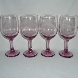 May include: Set of four clear glass wine glasses with a light pink tint. The glasses have a round bowl and a long stem.