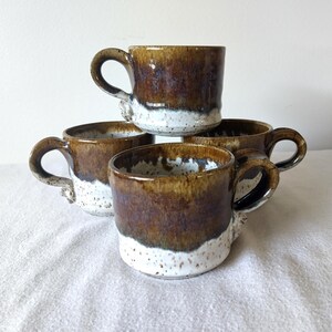 May include: Four handmade ceramic mugs. Each mug has a brown glazed top and a white speckled base. The mugs are stacked and arranged to show their unique shapes and textures. The mugs have large handles.