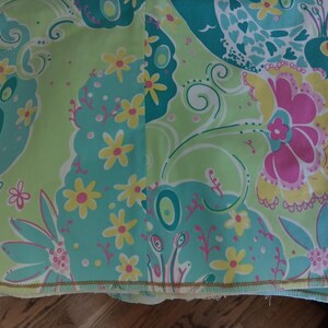 May include: A fabric textile featuring a repeating pattern of stylised peacocks, large floral blooms, and small yellow flowers. The colour palette consists of bright lime green, turquoise blue, hot pink, and yellow, with a finished hem along the bottom edge.