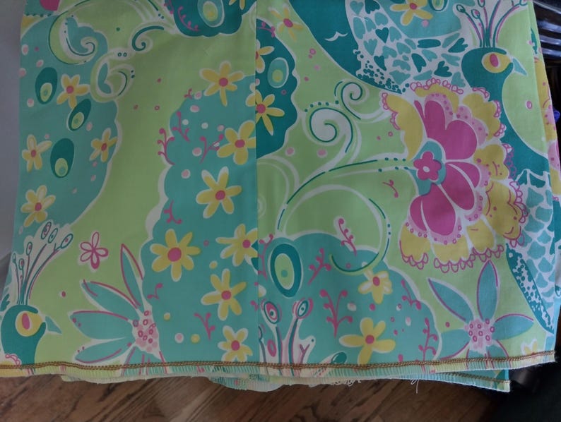 May include: A fabric textile featuring a repeating pattern of stylised peacocks, large floral blooms, and small yellow flowers. The colour palette consists of bright lime green, turquoise blue, hot pink, and yellow, with a finished hem along the bottom edge.
