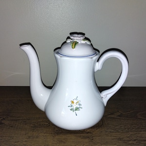 May include: White ceramic teapot with a curved spout and handle. The teapot features a floral design on the front and a flower-shaped knob on the lid. A blue line accents the rim of the lid. The teapot is sitting on a wooden surface.