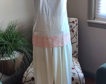 May include: A two-piece pajama set in cream-colored fabric featuring delicate peach-colored lace trim. The top is a camisole with thin straps and lace at the neckline and hem. The matching pants have a straight-leg cut with lace at the ankles.