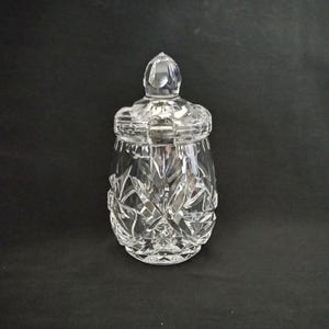 May include: A clear, cut-glass jar with a lid. The jar has a rounded shape with a textured, starburst pattern. The lid has a pointed finial. The jar is sitting on a black surface.