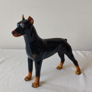 May include: A glossy ceramic figurine of a Doberman Pinscher dog standing in a profile pose. The sculpture features a black coat with tan markings on the muzzle, chest, and lower legs, with pointed ears and a smooth, reflective finish throughout.