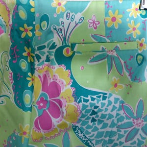 May include: A pair of shorts featuring a vibrant, multi-coloured pattern of stylised peacocks and flowers in shades of lime green, turquoise, bright pink, and yellow. The garment includes a single horizontal welt pocket on the right side.