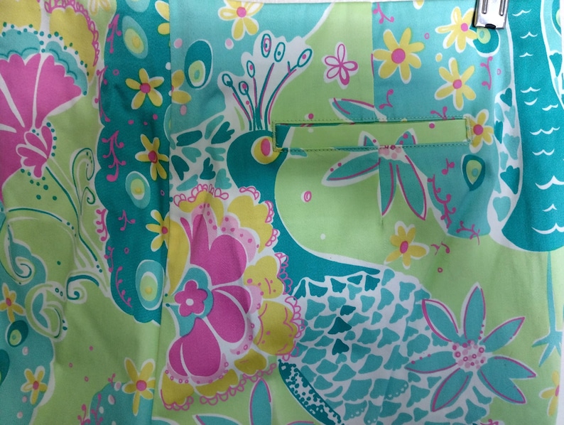 May include: A pair of shorts featuring a vibrant, multi-coloured pattern of stylised peacocks and flowers in shades of lime green, turquoise, bright pink, and yellow. The garment includes a single horizontal welt pocket on the right side.