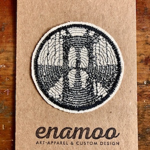 May include: A black and white embroidered patch with a circular design. The patch is on a brown card with the text "enamoo ART APPAREL & CUSTOM DESIGN".