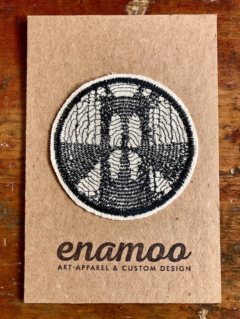 Może przedstawiać: Naszywka haftowana w kolorze czarno-białym z okrągłym wzorem. Naszywka znajduje się na brązowym kartonie z tekstem "enamoo ART APPAREL & CUSTOM DESIGN".