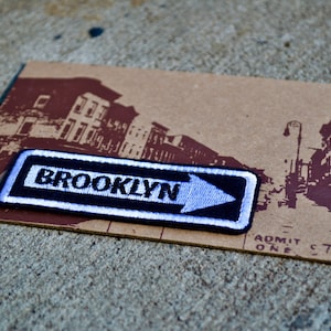 May include: A black and white fabric patch with a white arrow pointing to the right and the word "BROOKLYN" in white letters. The patch is on a brown background with a black and white image of a city street.