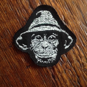 May include: A black embroidered patch of a monkey wearing a white hat. The monkey's face is white and the hat has a white brim.
