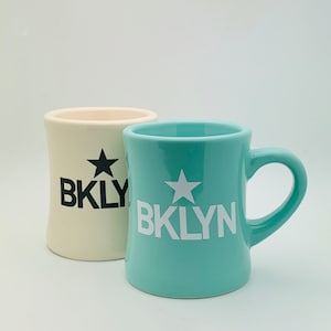 May include: Two ceramic mugs, one white with black lettering and a black star, and one turquoise with white lettering and a white star. Both mugs say "BKLYN".