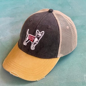 May include: A black and tan trucker hat with a mesh back. The hat features a small embroidered patch of a dog wearing a red vest with the word "Brooklyn" on it.