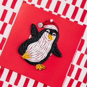 May include: A paper quilling art piece featuring a penguin wearing a red and white Santa hat. The penguin is crafted from coiled black, white, and yellow paper strips, displaying a textured, layered appearance on a solid red square background.