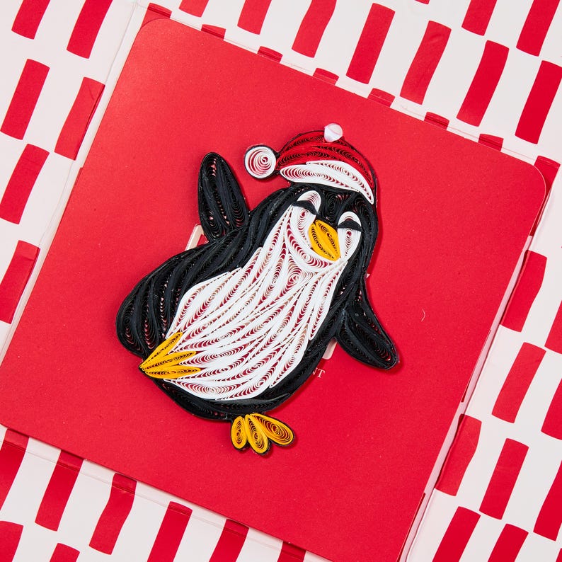 May include: A paper quilling art piece featuring a penguin wearing a red and white Santa hat. The penguin is crafted from coiled black, white, and yellow paper strips, displaying a textured, layered appearance on a solid red square background.
