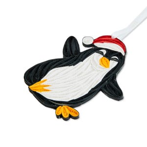 May include: A quilled paper ornament shaped like a penguin wearing a red and white Santa hat. The penguin is crafted from coiled black, white, and orange paper strips, featuring a white belly, black wings, and orange feet and beak.
