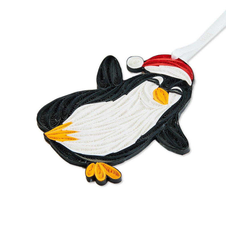 May include: A quilled paper ornament shaped like a penguin wearing a red and white Santa hat. The penguin is crafted from coiled black, white, and orange paper strips, featuring a white belly, black wings, and orange feet and beak.