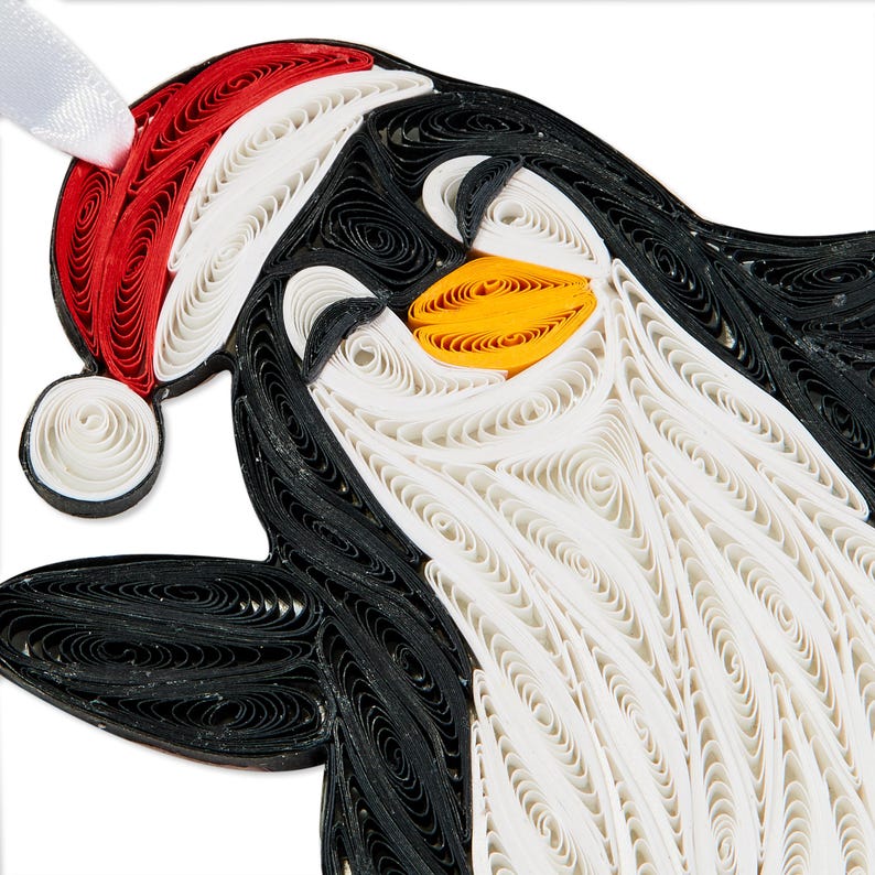 May include: A paper quilling ornament shaped like a penguin wearing a red and white Santa hat. The penguin is crafted from coiled strips of black, white, and orange paper, featuring a white ribbon loop at the top for hanging on a tree or display.