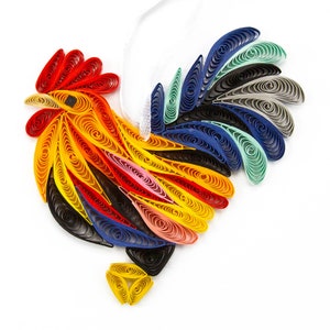 May include: A rooster-shaped ornament crafted from colorful paper quilling strips. The design features a red comb, a yellow head and body, and a tail composed of blue, teal, grey, and black coiled paper elements. A white ribbon loop is attached.