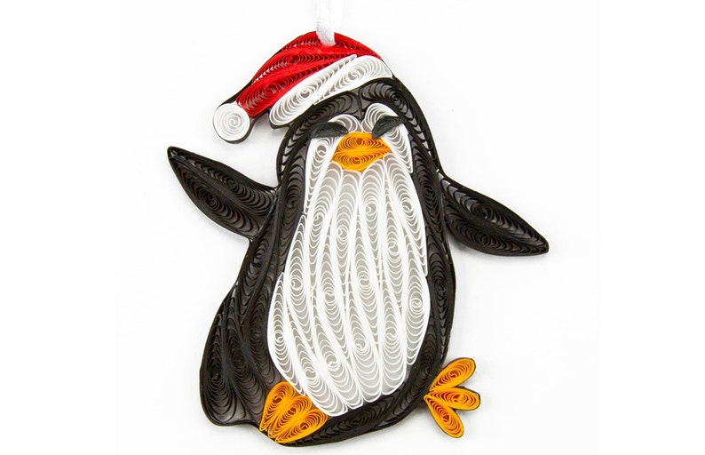 May include: A paper quilling ornament shaped like a penguin wearing a red and white Santa hat. The penguin is crafted from coiled black, white, and orange paper strips, featuring a white belly, orange feet and beak, and a white ribbon loop.