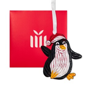 May include: A quilled paper penguin ornament featuring black, white, and orange coiled paper strips. The penguin wears a red and white quilled Santa hat and hangs from a white ribbon. A bright red square gift box with a white icon is behind it.