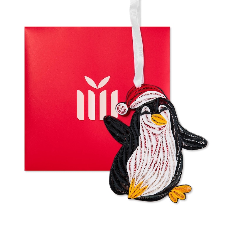 May include: A quilled paper penguin ornament featuring black, white, and orange coiled paper strips. The penguin wears a red and white quilled Santa hat and hangs from a white ribbon. A bright red square gift box with a white icon is behind it.