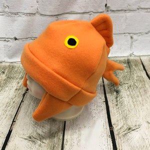 May include: An orange fleece hat shaped like a fish with a black eye and yellow outline. The hat has ear flaps and a fin on the back.