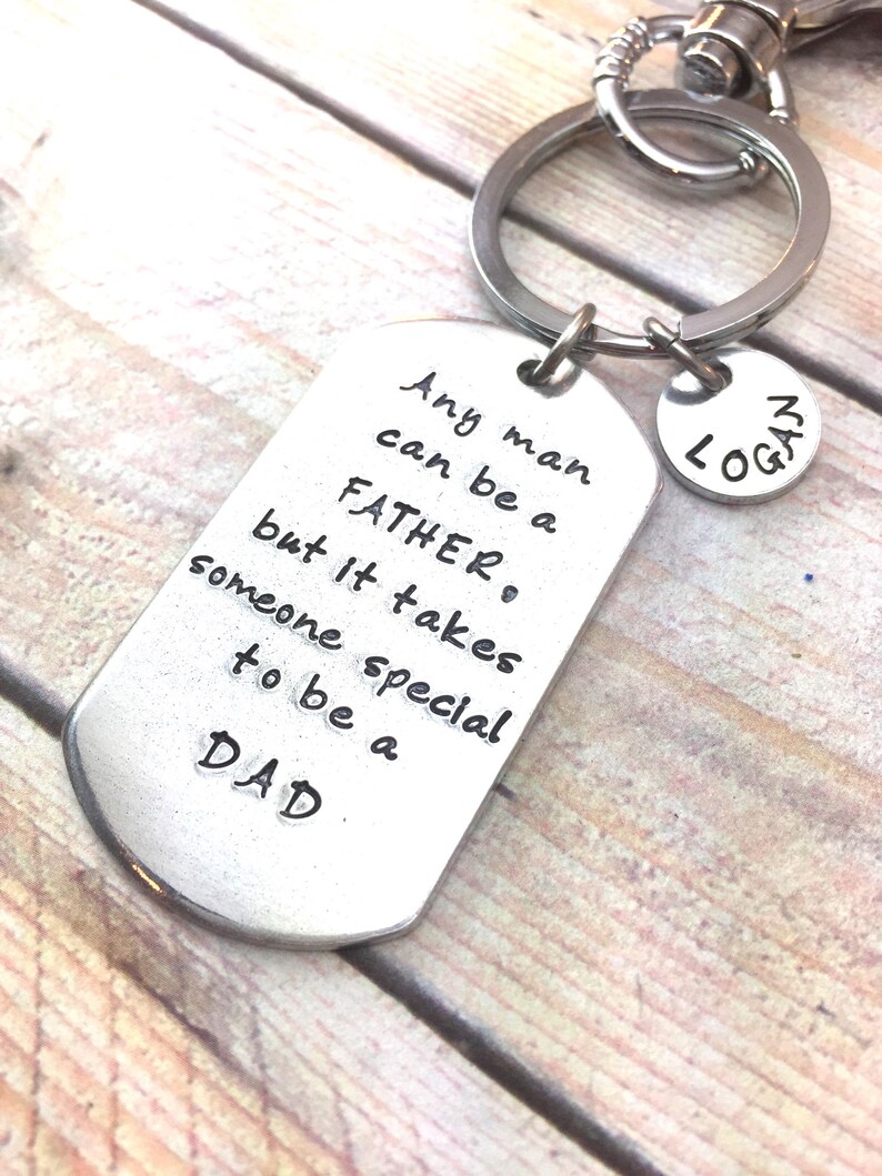May include: Silver keychain with a dog tag charm that reads "Any man can be a father, but it takes someone special to be a dad."  A smaller silver charm with the name "Logan" is attached to the keychain.