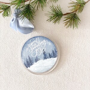 May include: A round, white ornament with a winter scene and the text "repeat the sound of joy" in white calligraphy.