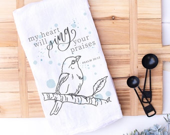 May include: White kitchen towel with a blue watercolor design and a black and white illustration of a bird on a branch. The text "my heart will sing your praises" is written in black cursive script. The text "PSALM 30:12" is written in smaller black font.