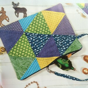 Könnte beinhalten: Eine bunte Patchwork-Stofftasche mit Reißverschluss und Trageriemen. Die Tasche besteht aus verschiedenen gemusterten Stoffen in Grün-, Blau-, Gelb- und Lilatönen. Die Tasche liegt auf einer weißen Oberfläche mit einer goldenen Kette und einem grünen Ornament.