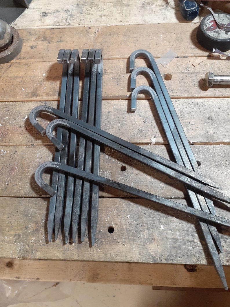 May include: A set of ten metal tent pegs with a hooked end. The pegs are arranged in a criss-cross pattern on a wooden surface.