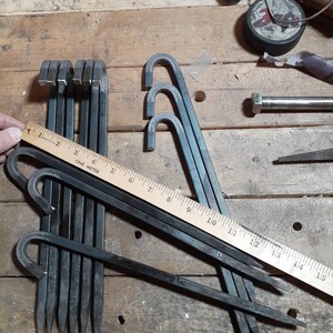 May include: A set of nine black metal tent pegs with a hook on one end. The pegs are arranged in a pile on a wooden surface. A ruler is visible in the image, showing the pegs are approximately 38 cm long.
