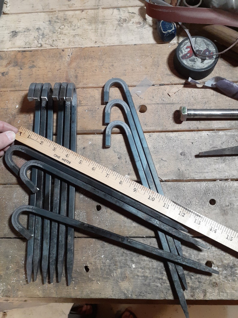 May include: A set of nine black metal tent pegs with a hook on one end. The pegs are arranged in a pile on a wooden surface. A ruler is visible in the image, showing the pegs are approximately 38 cm long.