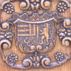 Könnte beinhalten: Eine hellbraune Holzplatte mit einer komplexen Reliefschnitzerei eines heraldischen Schildes. Der Schild zeigt einen Löwen, horizontale Balken und ein ankerartiges Symbol. Das Design ist von kunstvollen Blumen- und Rankenmustern umgeben.