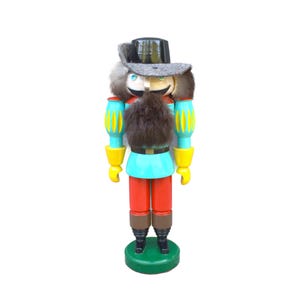 May include: A colorful wooden nutcracker figurine. It features a black top hat, a gray brim, and a faux fur beard. The figurine has a turquoise shirt, red pants, and yellow accents. It stands on a green base.