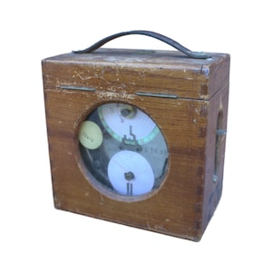 Op de afbeelding: Een vintage houten doos met een donkerlederen handvat aan de bovenkant en een rond glazen venster aan de voorzijde. Binnenin zijn twee witte ronde papieren schijven zichtbaar, samen met een gele sticker met het nummer 02413 en 61690.