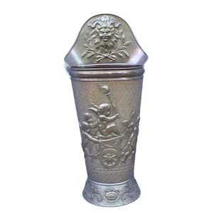 May include: A bronze-colored umbrella stand with classical relief carvings. The stand features a decorative top with a face and laurel wreath design. The body of the stand is adorned with cherubs and a chariot scene.