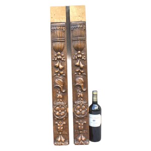 May include: Two tall, rectangular wooden architectural panels featuring intricate relief carvings of urns, floral motifs, and helmet shapes. The wood has a medium brown finish. A dark glass wine bottle stands next to the panels for scale.